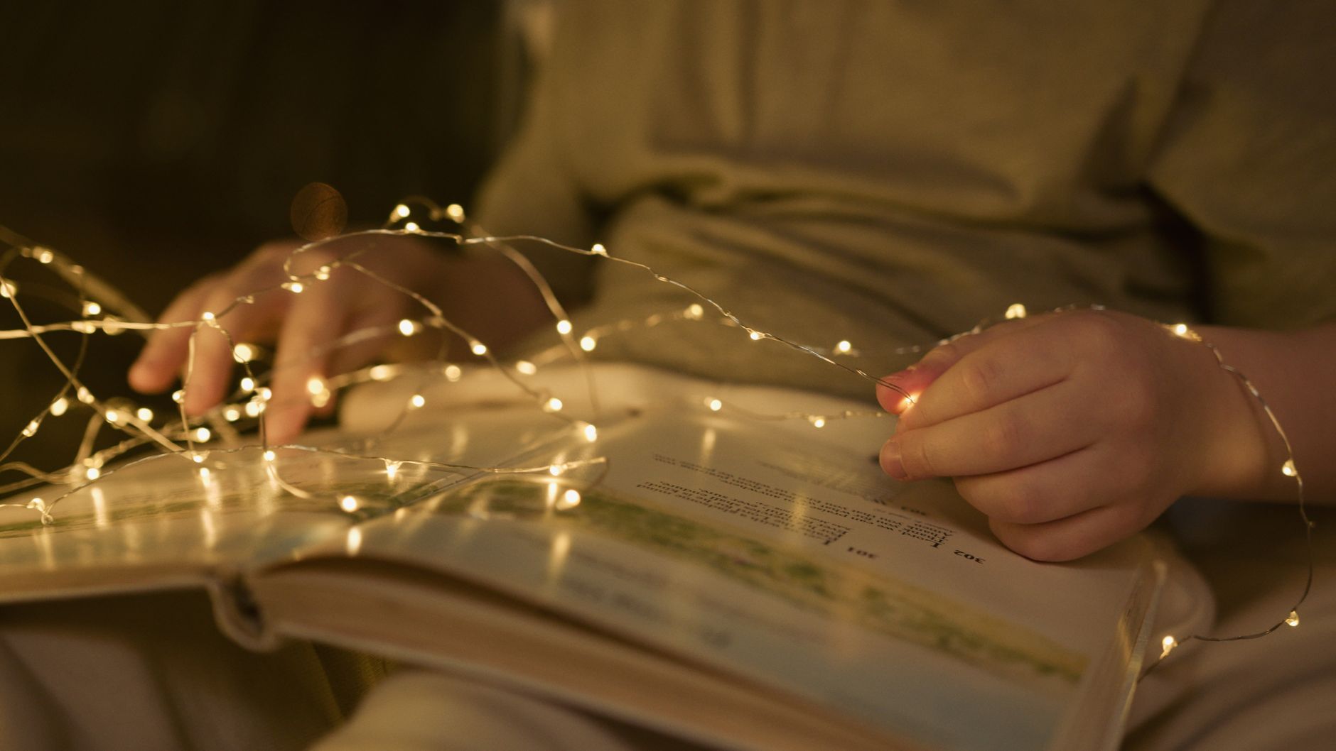 Открытая иллюстрированная книга в руках ребёнка, рядом тёплая гирлянда